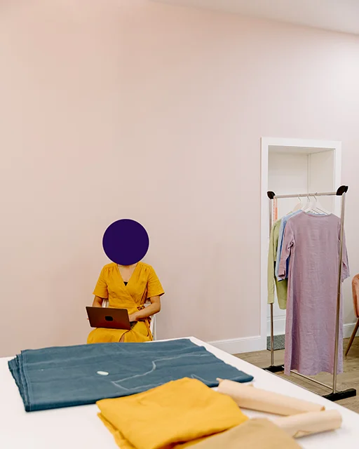 A person in a yellow dress working on a laptop in a bright studio, with hanging garments nearby