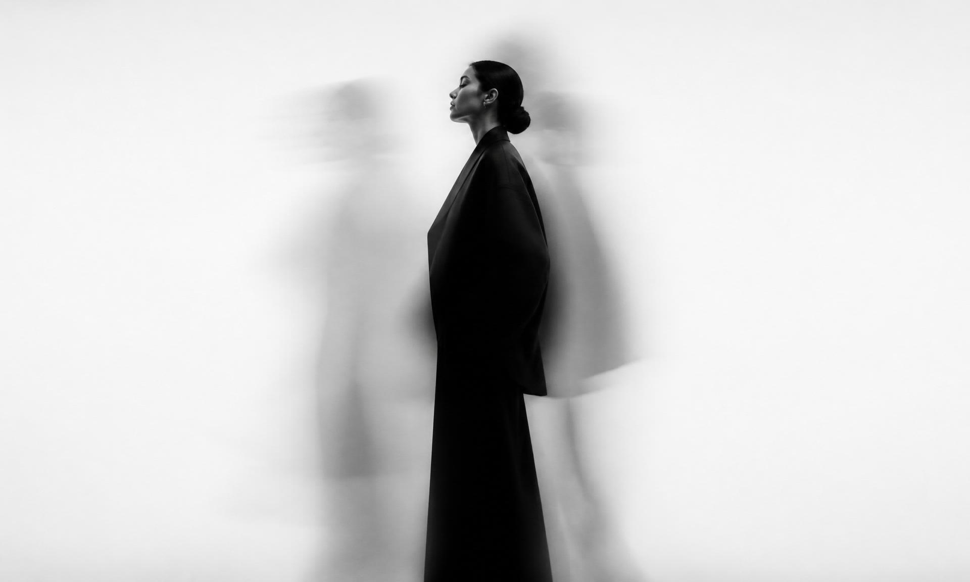 A woman in dark minimal clothing stands still against a white background, blurred figures passing behind her — quiet restraint in motion